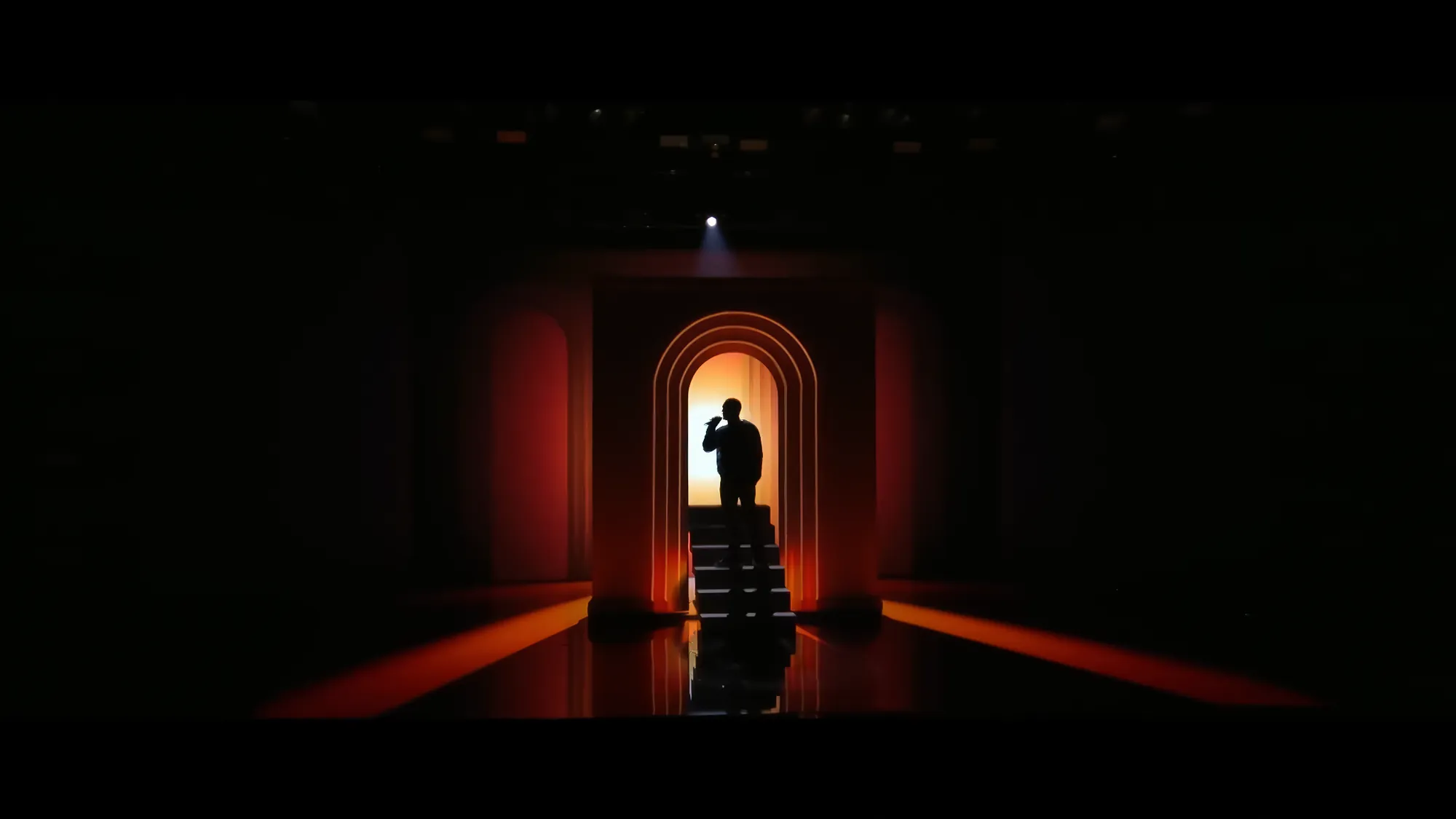 The darkened stage, a lone figure in silhouette at the top of a staircase inside a tall arch of deep red light, reflected in the floor