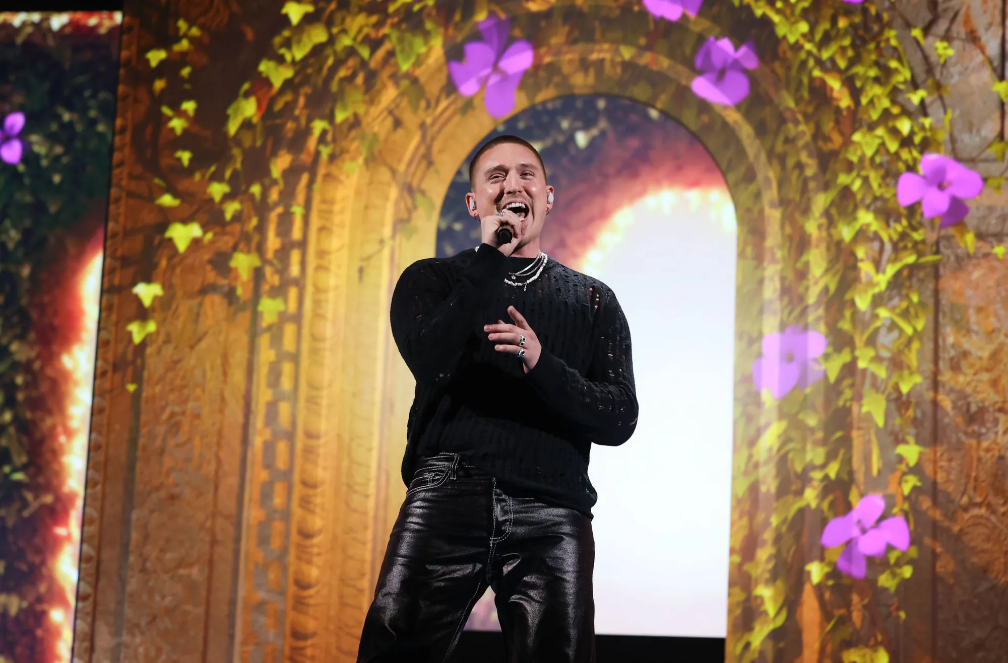Danny Saucedo singing in front of the gate, its arch overgrown with projected ivy and purple flowers in warm gold light