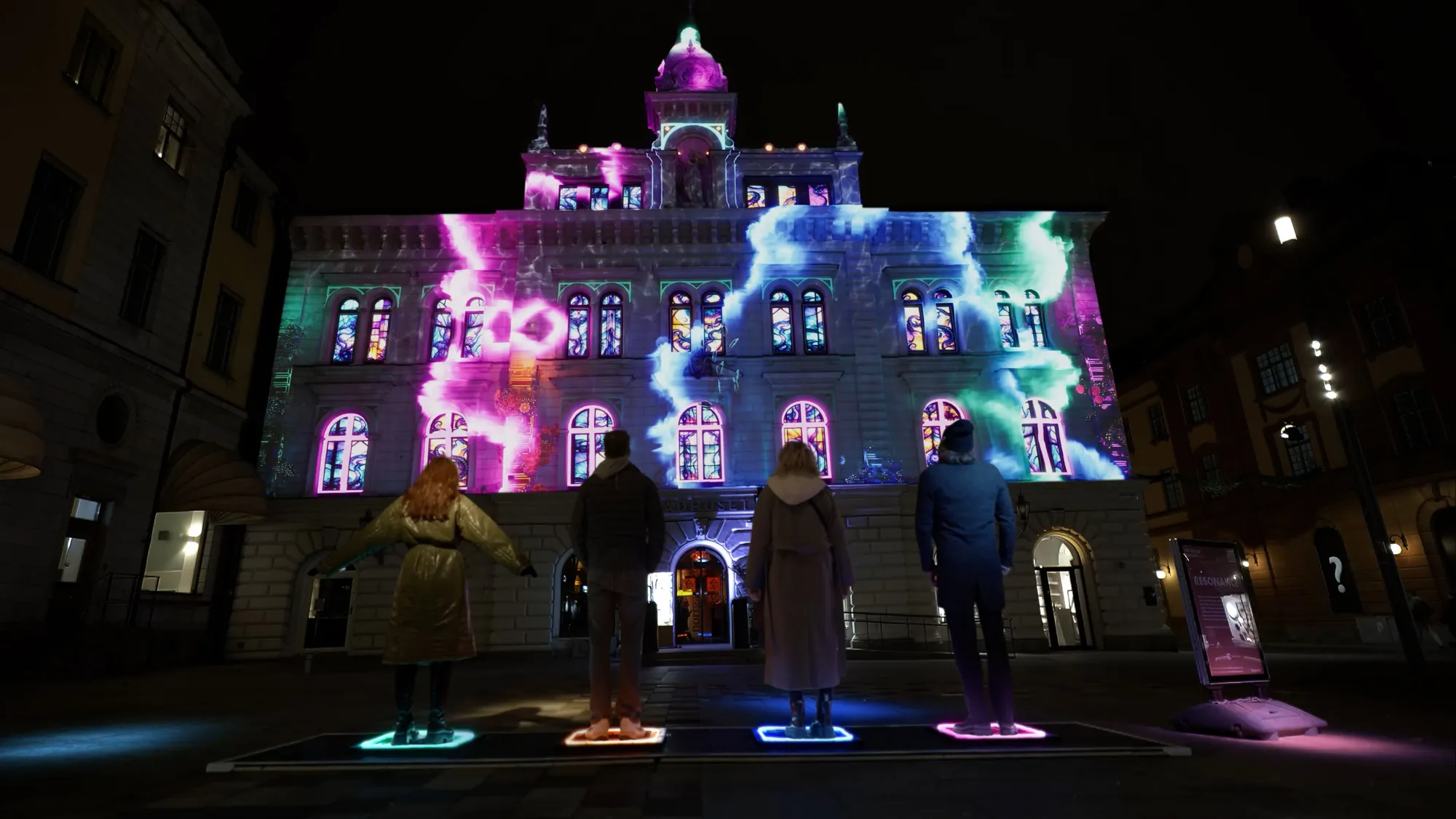 Uppsala Town Hall at night, its whole facade washed in pink and blue projection, people standing on lit pads in the square below