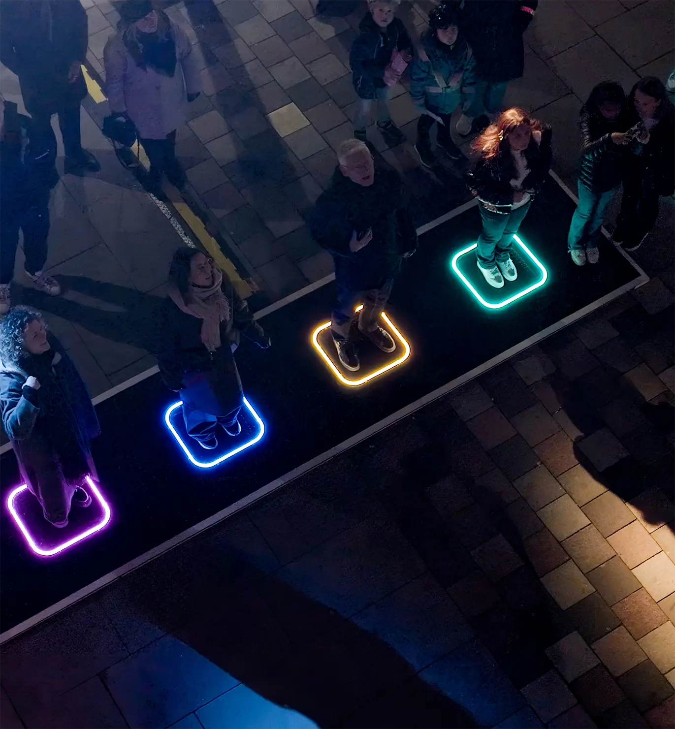 The four touch pads seen from above, glowing green, blue, amber and teal on the wet cobbles with people gathered around them