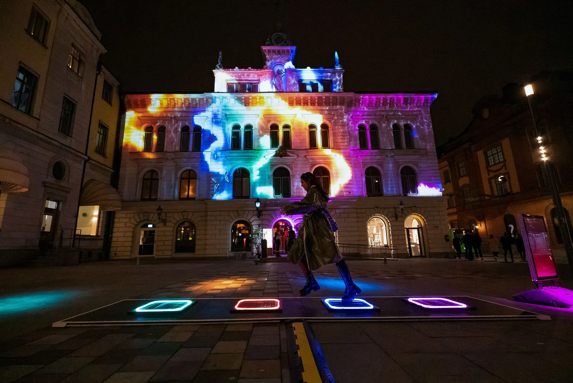 The four touch pads seen from above, glowing green, blue, amber and teal on the wet cobbles with people gathered around them